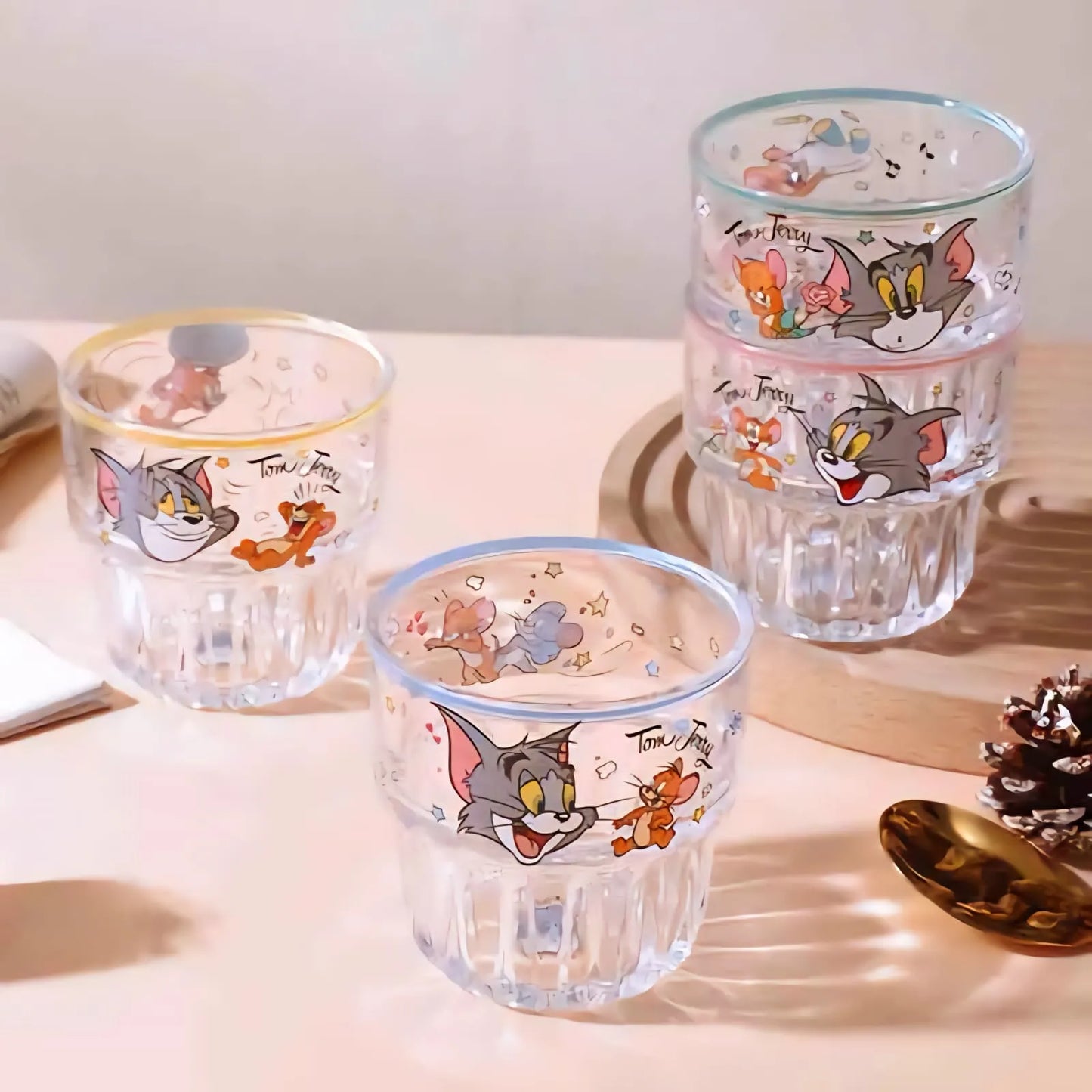 Three clear ribbed drinking glasses with cartoon Tom and Jerry prints, arranged on a light tabletop with decorative props.