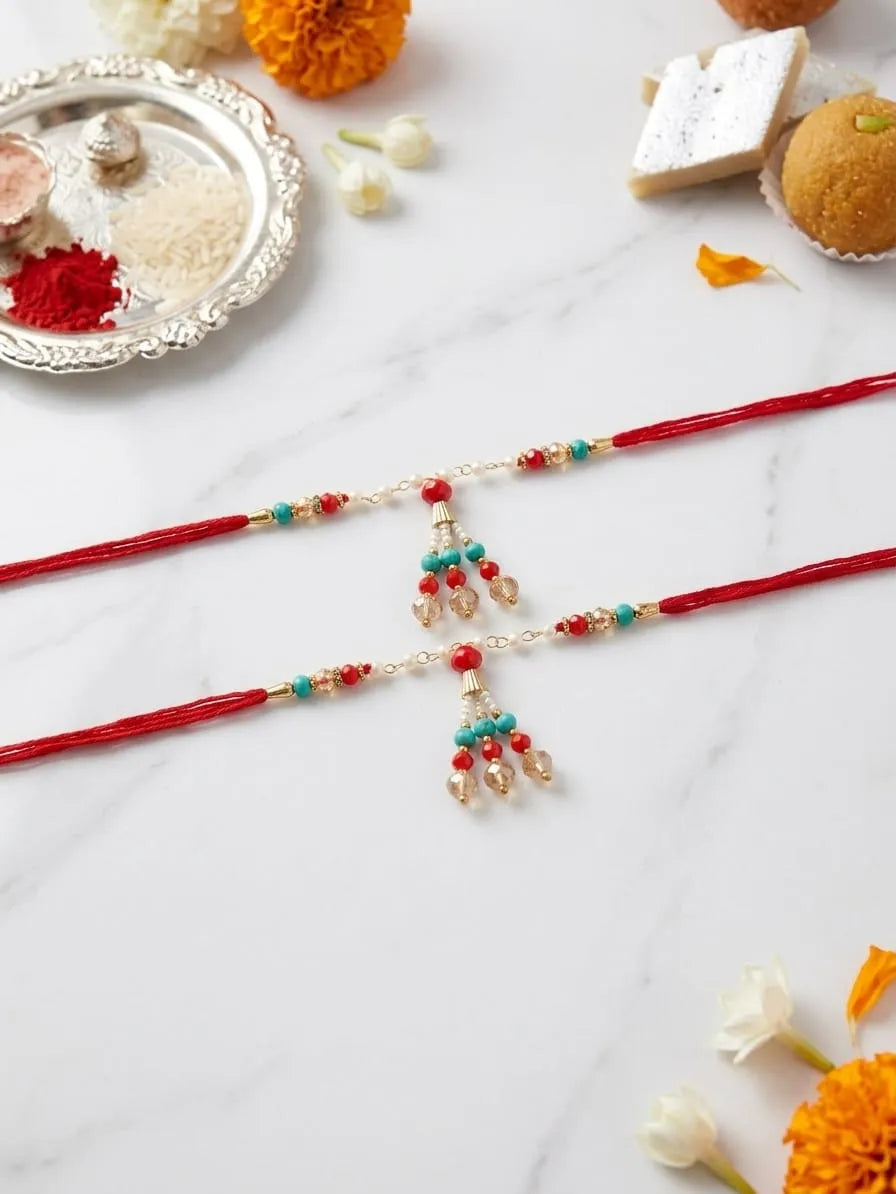 Two red Rakhi cords laid flat on a white marble surface, decorated with small gold beads, turquoise and red bangles, and dangling gold-toned charms. Decorative sweets, flowers, and a silver plate are arranged around them.