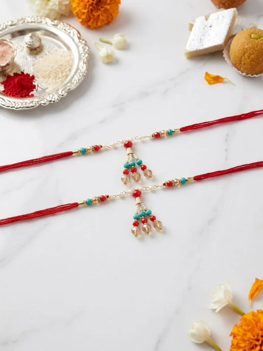 Two red Rakhi cords laid flat on a white marble surface, decorated with small gold beads, turquoise and red bangles, and dangling gold-toned charms. Decorative sweets, flowers, and a silver plate are arranged around them.