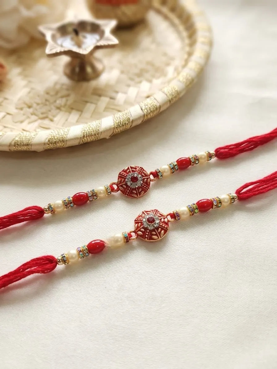 Two red and gold beaded Rakhi bracelets laid on a light cream fabric surface, with a woven tray and diya in the softly blurred background.