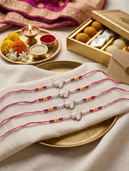 Four Rakhi string bracelets with white elephant charms and colorful beads are arranged on a cream-colored fabric over a gold plate, with a traditional Diwali gift box and lit diya in the background.