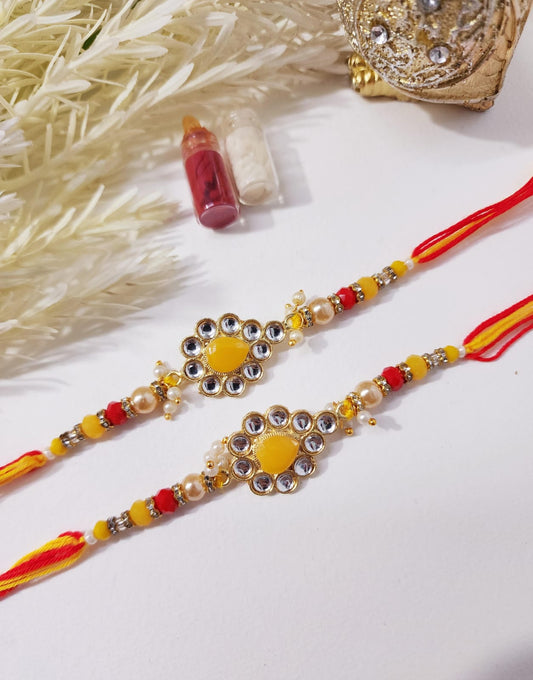 Two HOK Yellow Kundan Rakhi with intricate design and red and yellow color scheme, displayed with decorative items in the background.