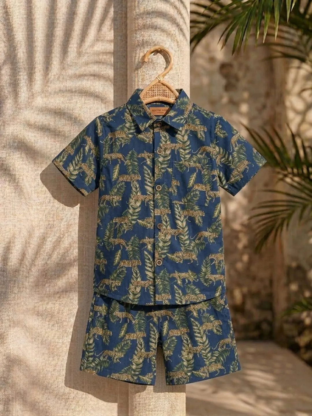 Blue and green patterned shirt and shorts set on a hanger against a neutral background with plants.