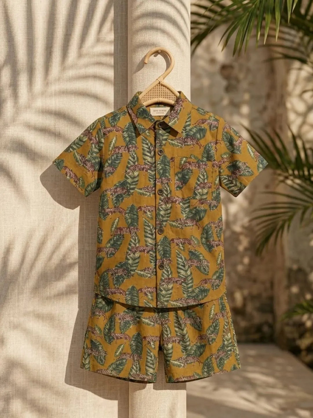 Tropical patterned shirt and shorts set hanging on a wooden hanger against a neutral background with palm leaves.