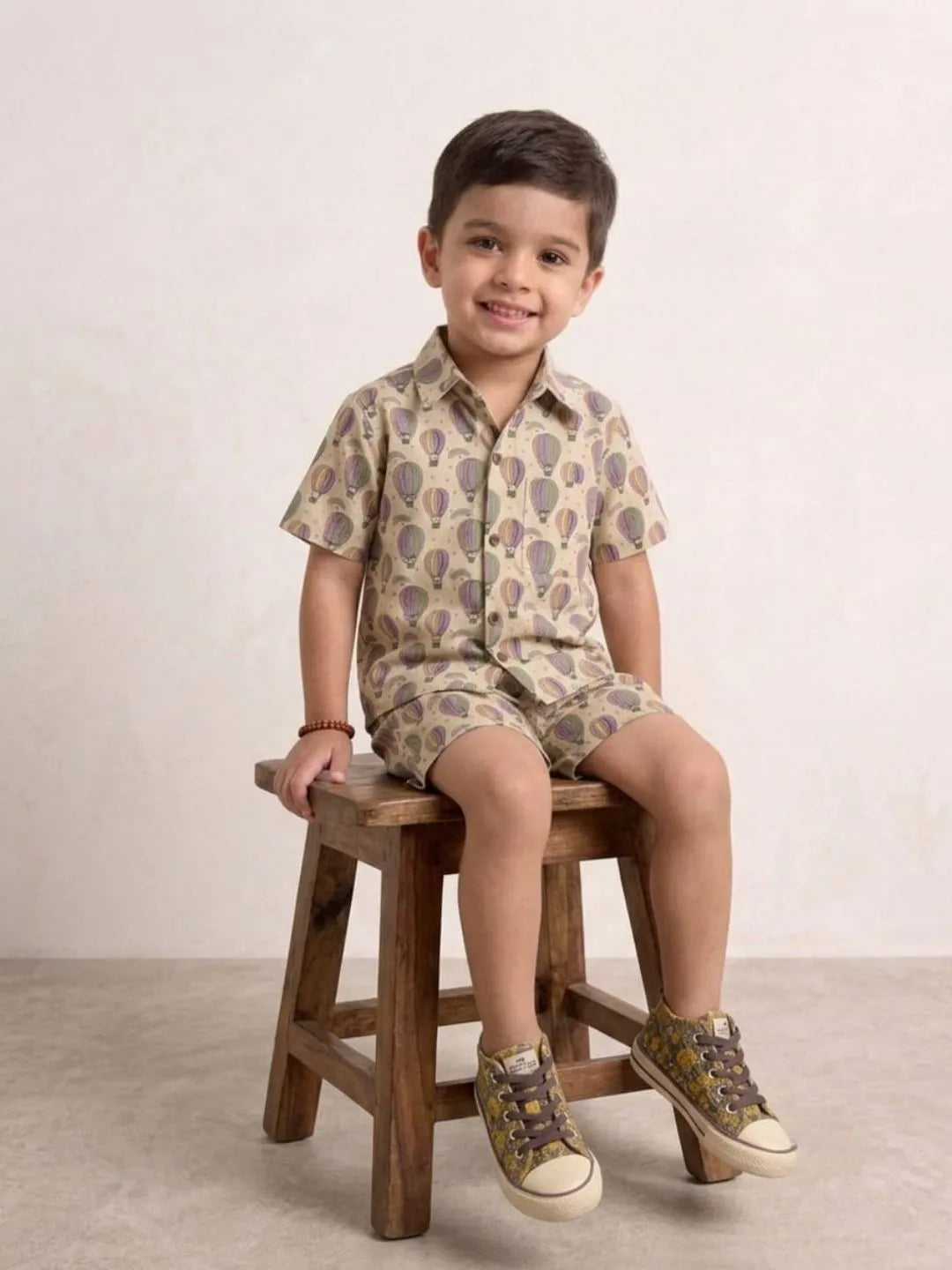 Child sitting on a wooden stool wearing a patterned shirt and shorts set.
