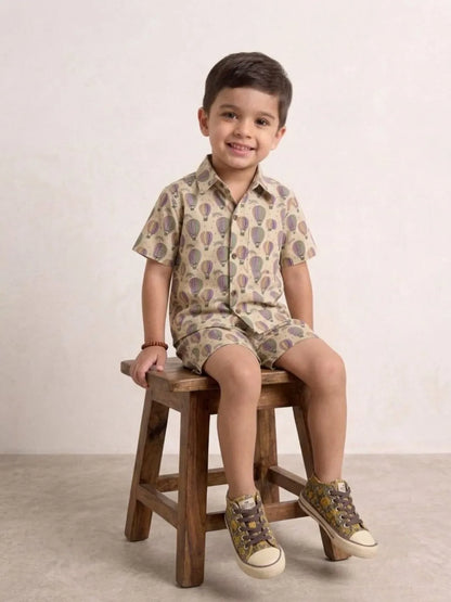 Child sitting on a wooden stool wearing a patterned shirt and shorts set.
