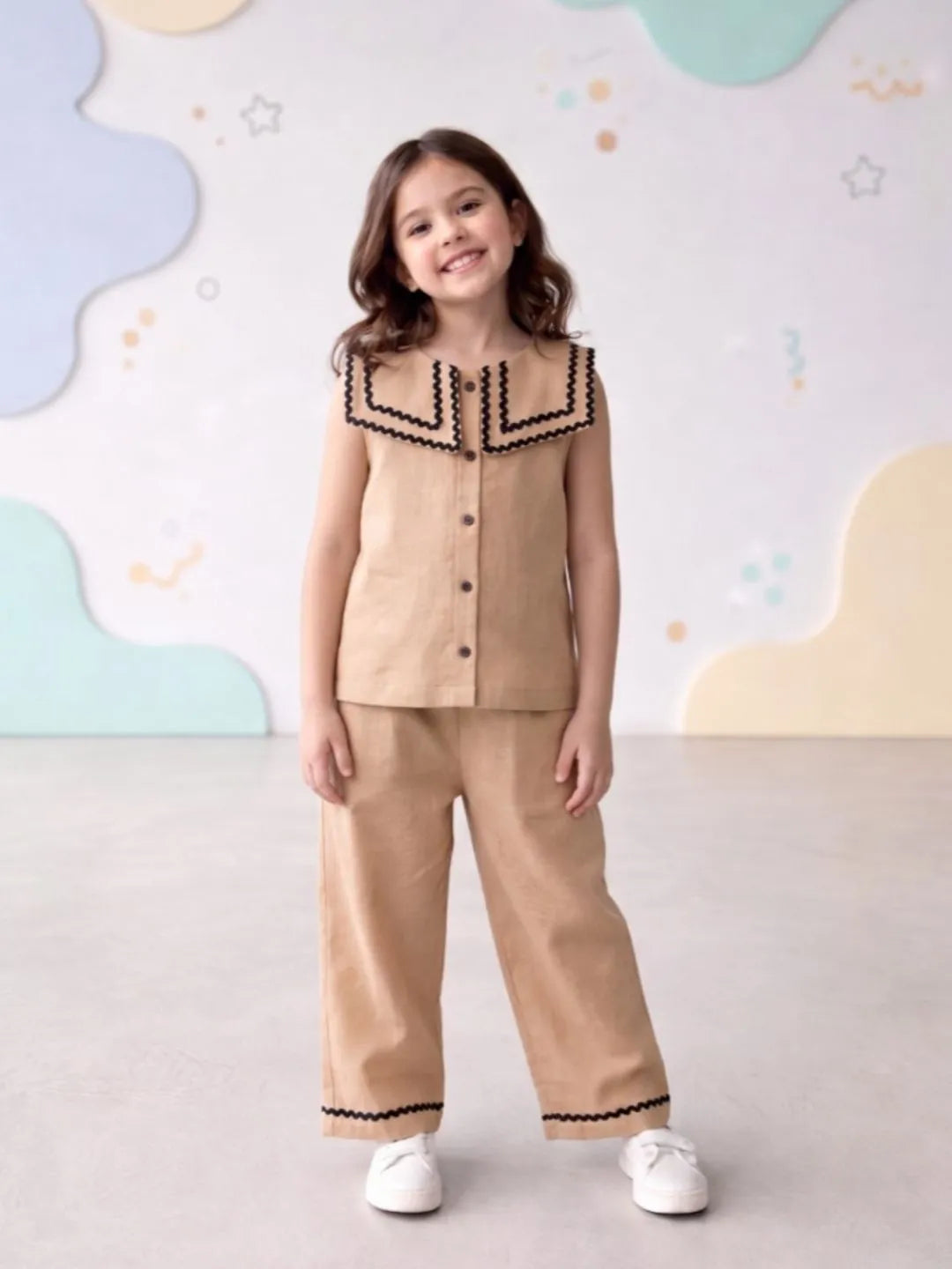 Young girl wearing a beige outfit with black trim against a colorful wall.