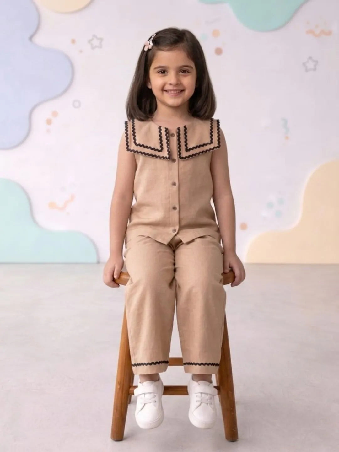 Young girl in a beige outfit with black trim sitting on a wooden stool against a colorful wall.