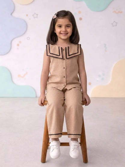Young girl in a beige outfit with black trim sitting on a wooden stool against a colorful wall.
