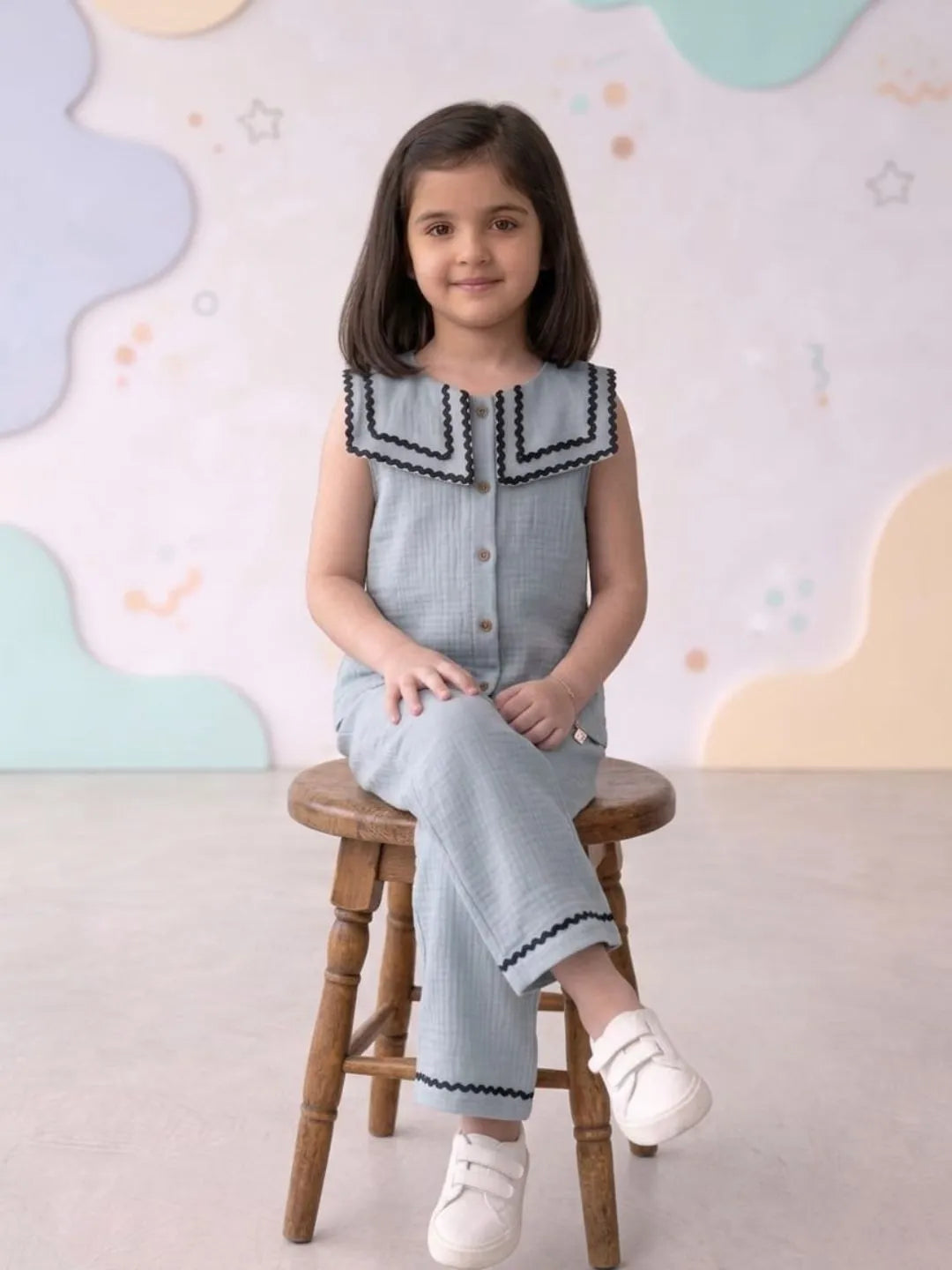 Young girl sitting on a wooden stool wearing a light blue dress with a dark trim against a colorful cloud-patterned wall.