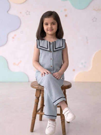 Young girl sitting on a wooden stool wearing a light blue dress with a dark trim against a colorful cloud-patterned wall.