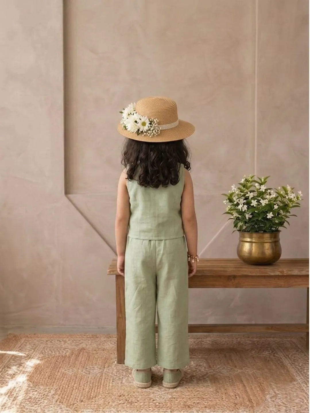 Child in a green jumpsuit and straw hat standing in front of a beige wall with a wooden bench and potted plant.