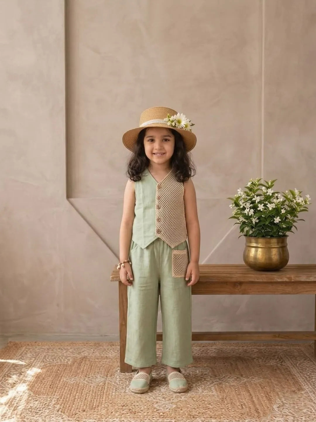 Young girl in a green outfit with a hat standing in front of a beige wall with a wooden bench and flowers.