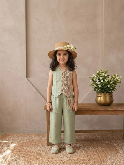 Young girl in a green outfit with a hat standing in front of a beige wall with a wooden bench and flowers.