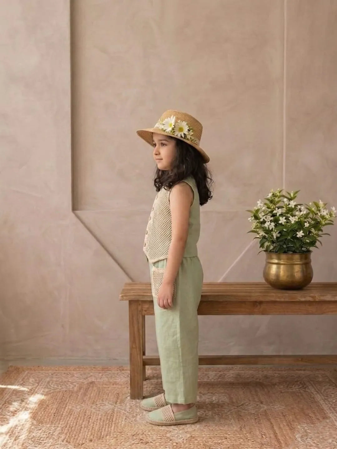 Young girl in a beige hat and green outfit standing in a room with a wooden bench and potted plant.