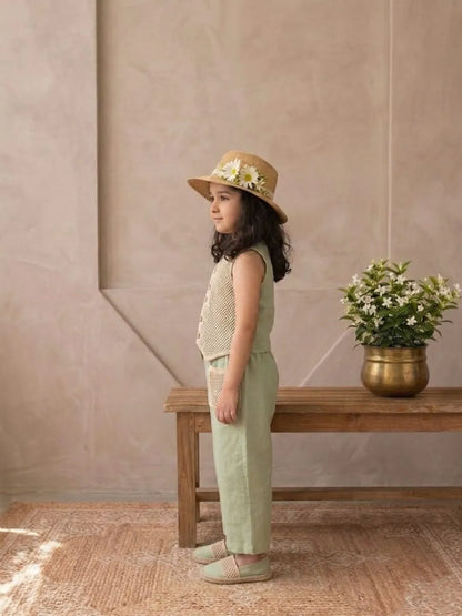 Young girl in a beige hat and green outfit standing in a room with a wooden bench and potted plant.