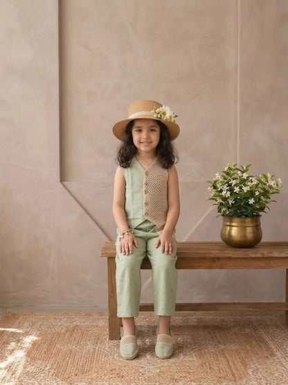 Young girl in a green outfit and hat sitting on a wooden bench with a plant in the background.