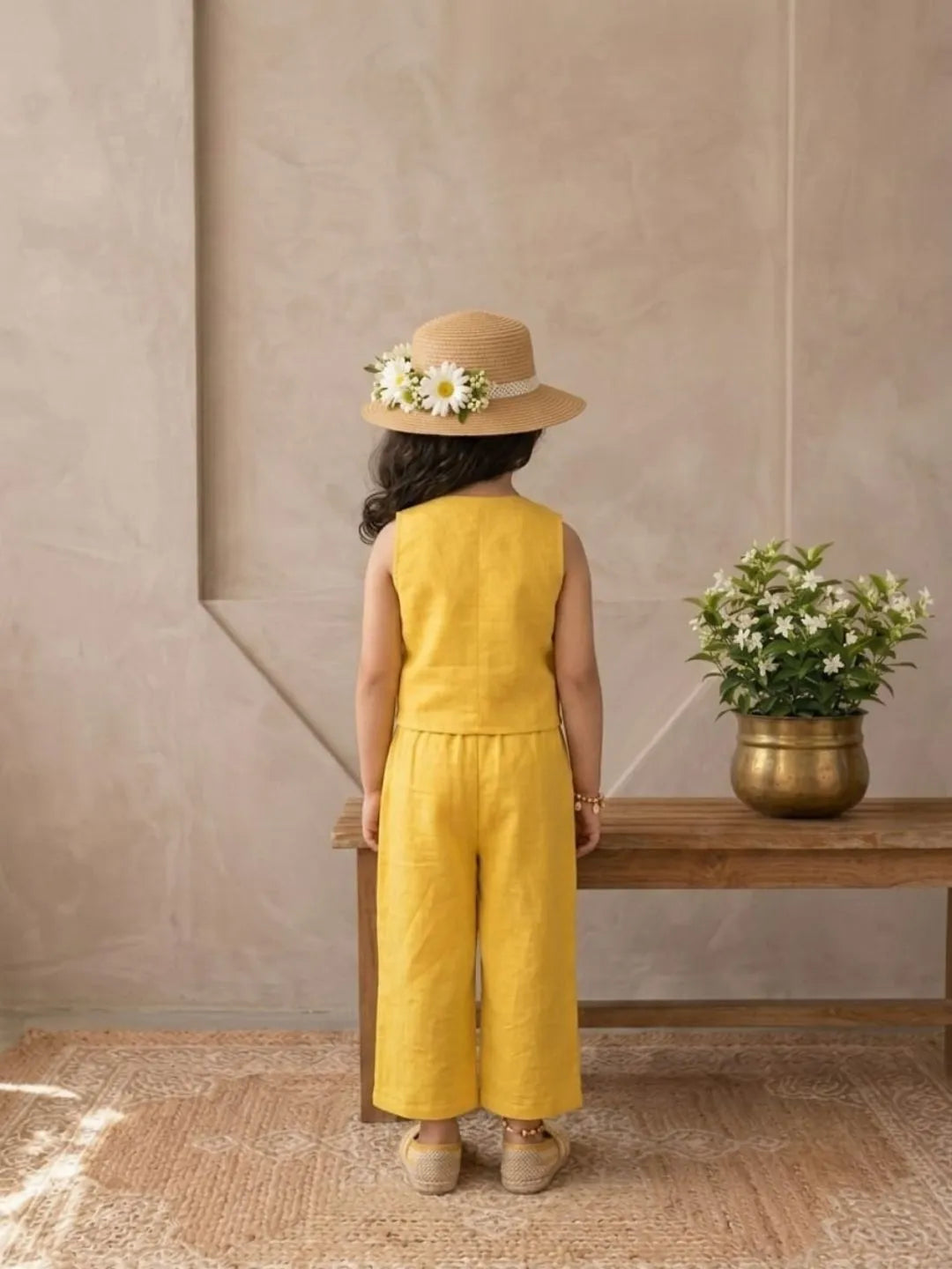 Child in a yellow outfit and hat standing near a wooden bench with a plant, against a beige wall.