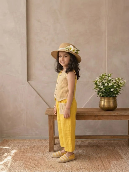 Young girl in yellow outfit with hat standing next to a wooden bench with a plant on it.