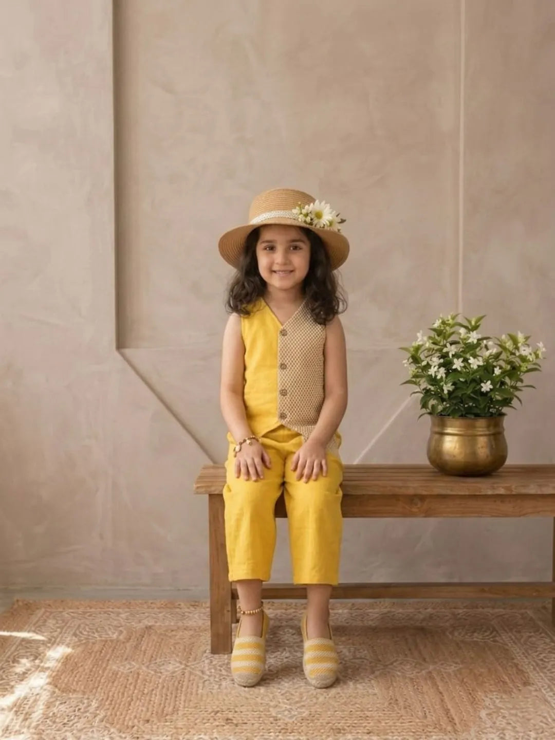 Young girl in a yellow outfit with a hat sitting on a wooden bench against a beige wall.