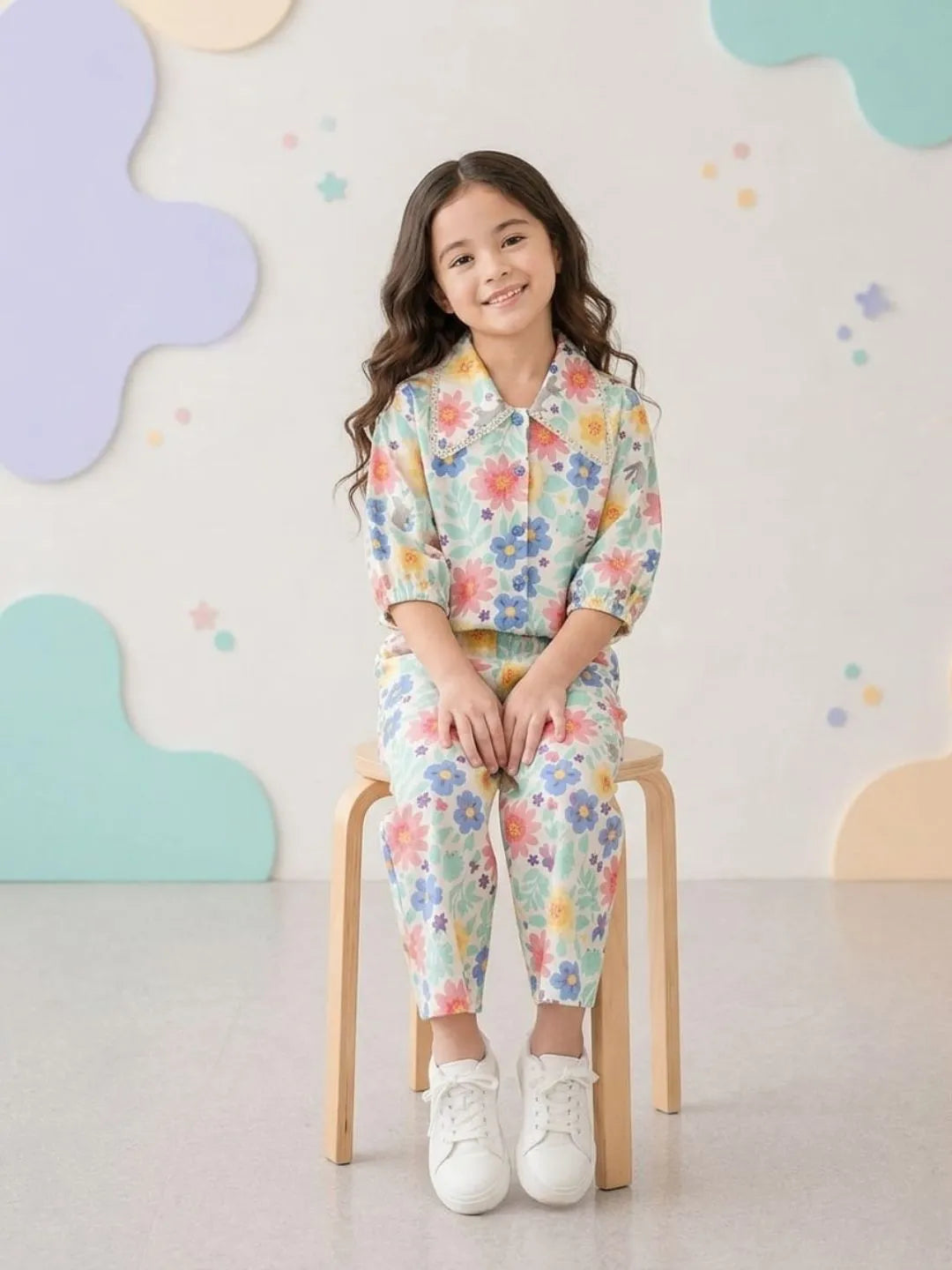 Young girl sitting on a chair wearing a floral outfit with a colorful cloud-patterned wall in the background.